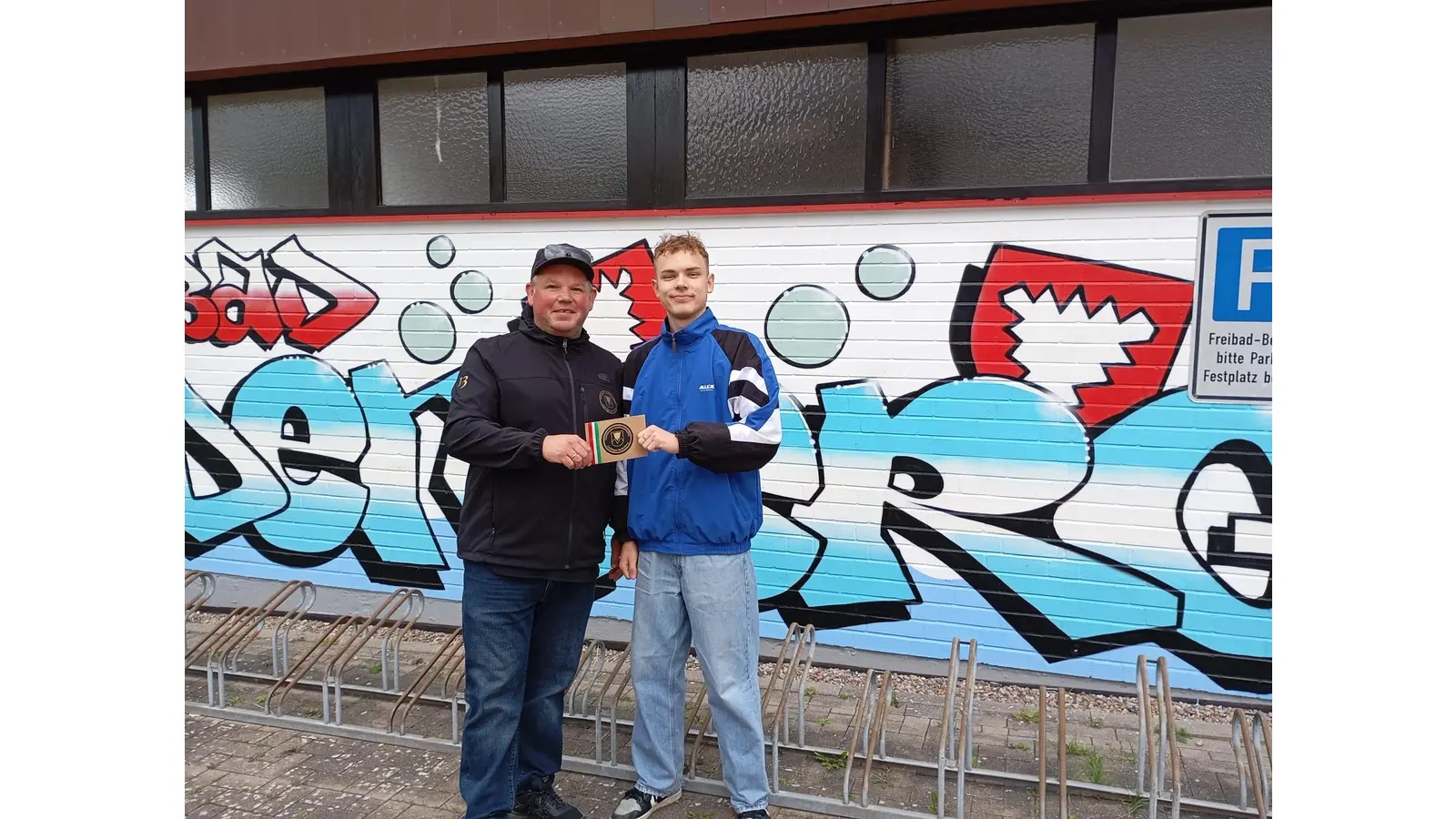 Steffen Buller von der Interessengemeinschaft Freibad Rodenberg übergibt eine kleine Geldspende an Leo (19) den Schöpfer des Graffitis.  (Foto: privat)