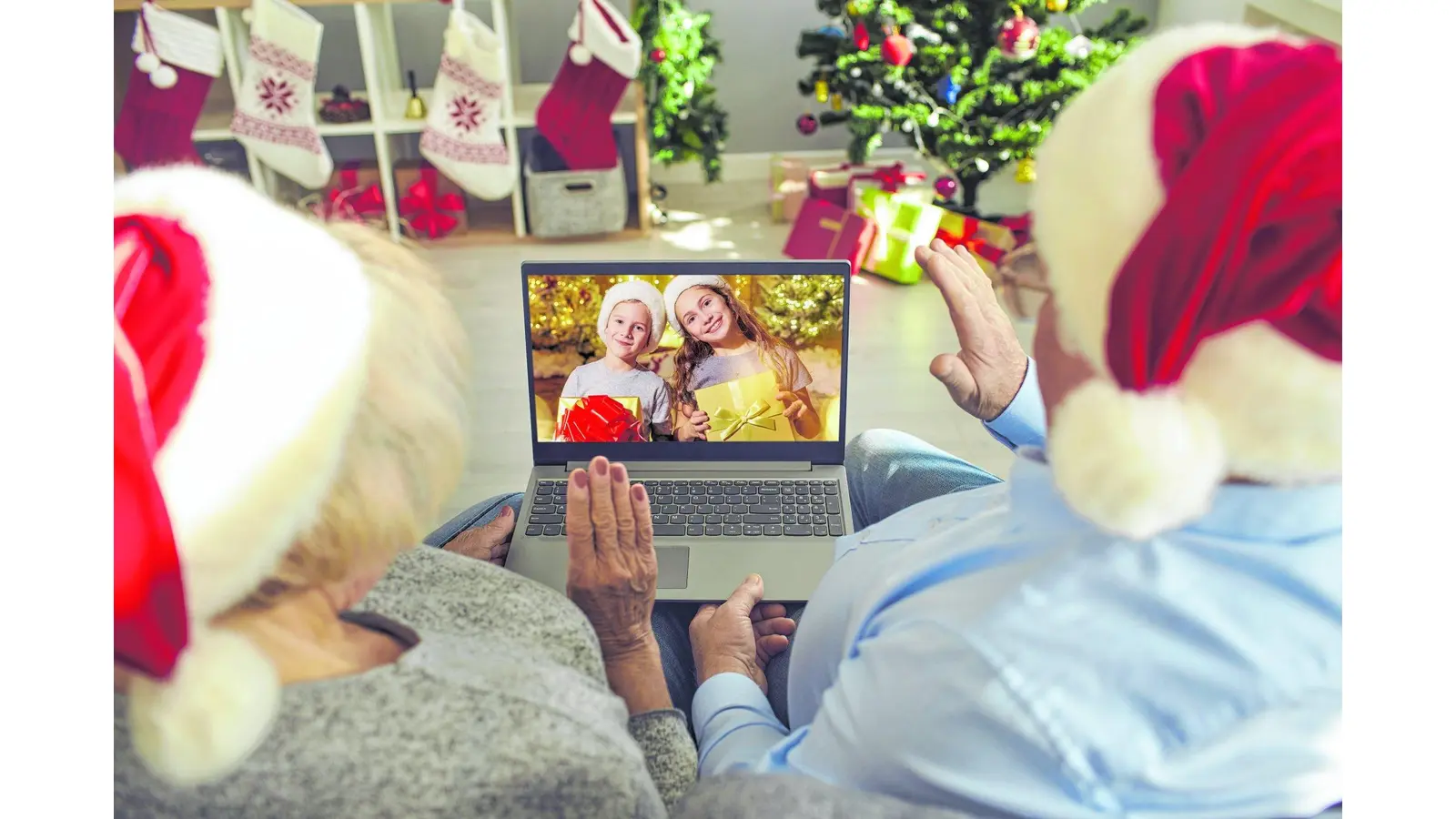 Staying at home during quarantine, Christmas in self-isolation, virtual family meeting. Happy grandparents in Santa hats sitting on sofa together video calling grandchildren. Back view over shoulder (Foto: mk)